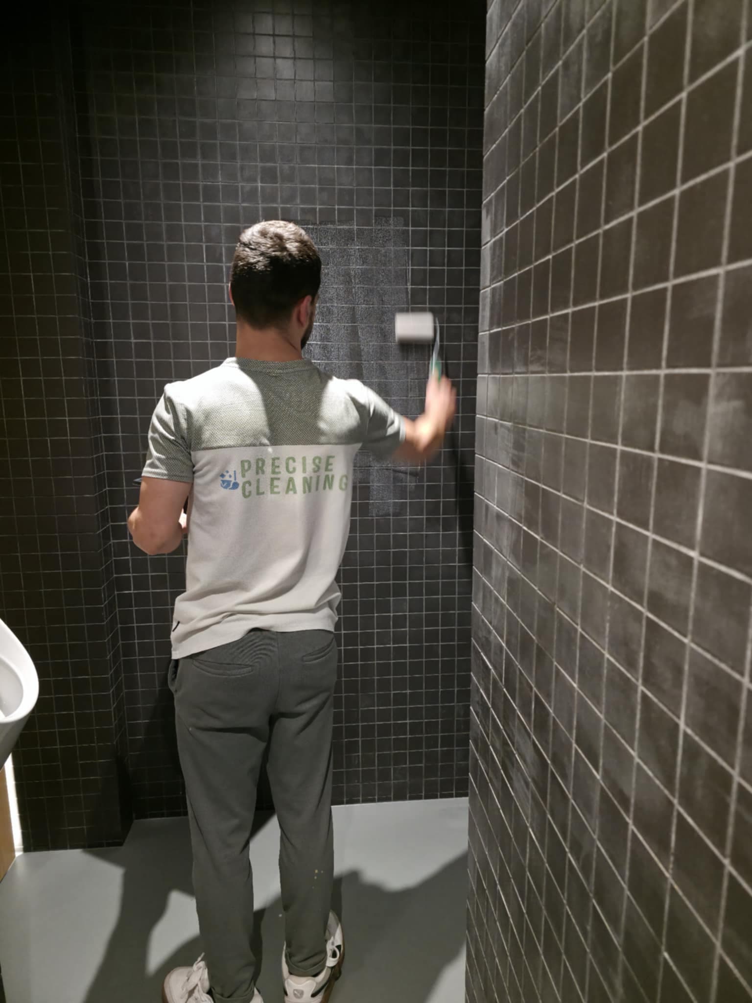 Man wearing a Precise Cleaning shirt uses a roller on dark grey square tiled walls.