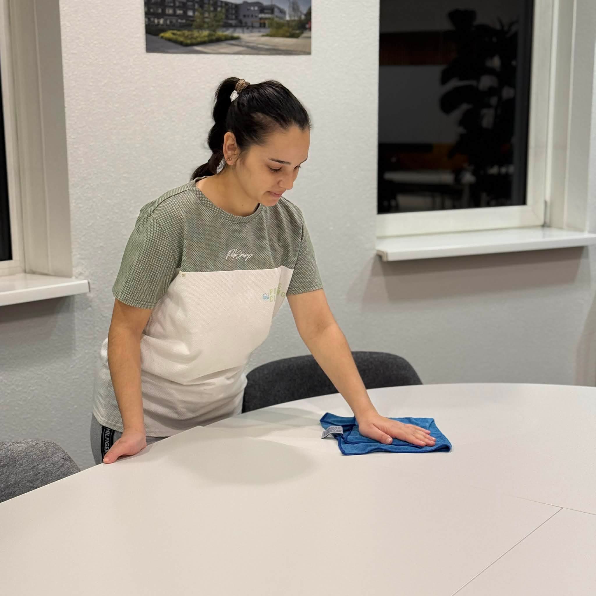 Woman with a ponytail wipes a large white table using a blue microfiber cloth.