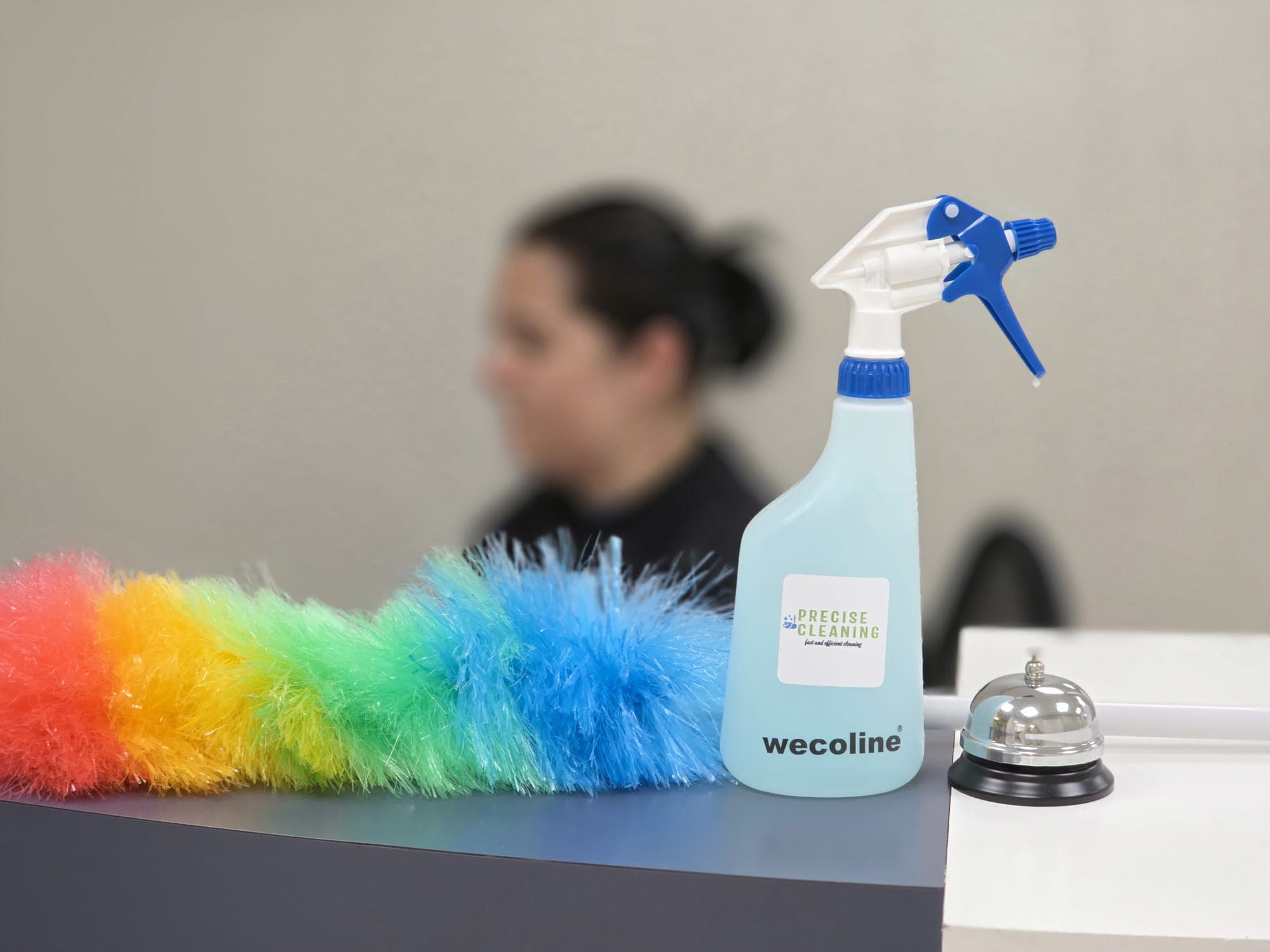 Rainbow duster, blue spray bottle, and silver service bell on a counter with blurred background.